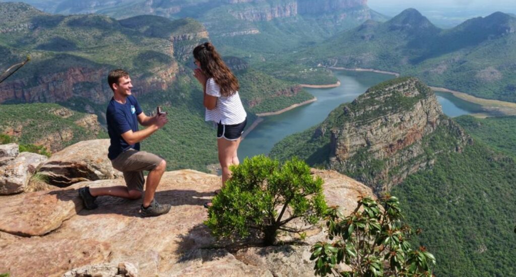 An image of a man on his knees proposing to his girlfriend on a mountain-like area. Image Source: Unknown