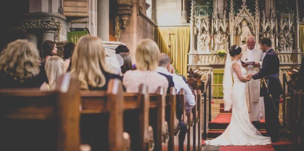 A couple pictured exchanging wedding vows with a priest as an officiator. Image Source: Getty