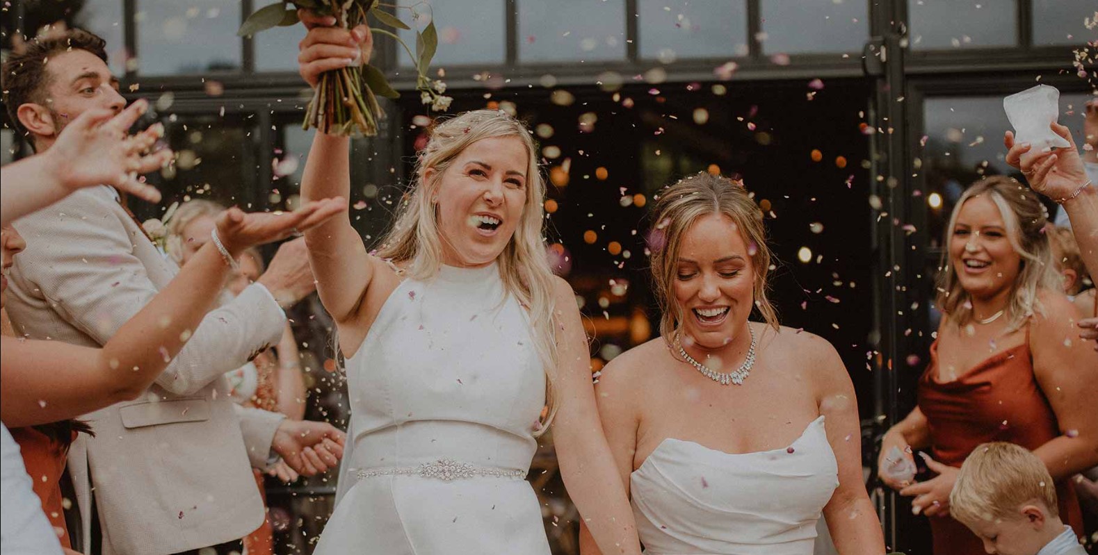 Two women celebrating at their LGBTQI+ wedding in the North East of England. They are both holding flowers and hands, walking through their guests laughing while everybody throws confetti. Image Source: iStock Photos